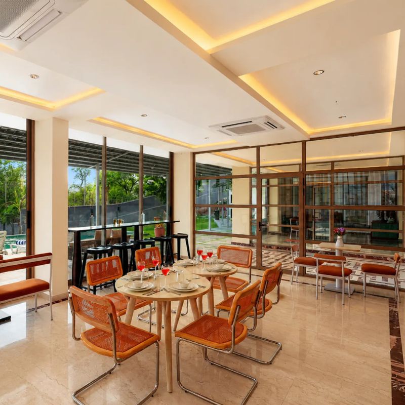 Indoor dining table set for guests with orange chairs and pool views at a 9 BHK villa Udaipur.